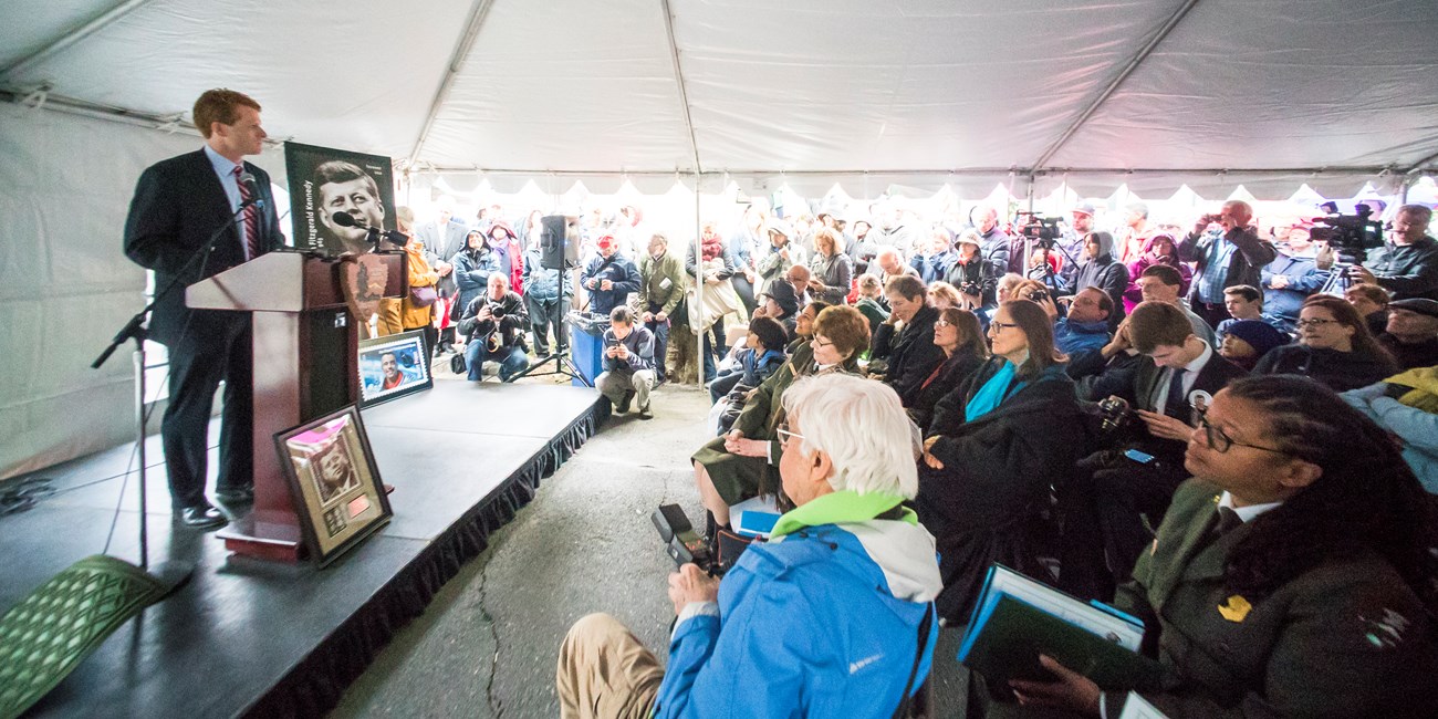 Joseph Kennedy III is dressed in a suit and standing on a stage behind a podium.  A large crowd is gather under a white tent.