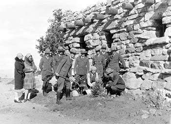 A group or people are shown planting a tree in front of a stone house.  All of the men are uniformed rangers.  Two women in long coats can be seen standing apart from the group.