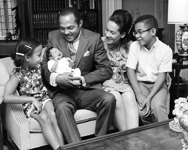 An African American man sits on a sofa with this wife and two older children. He holds an infant in his arms.