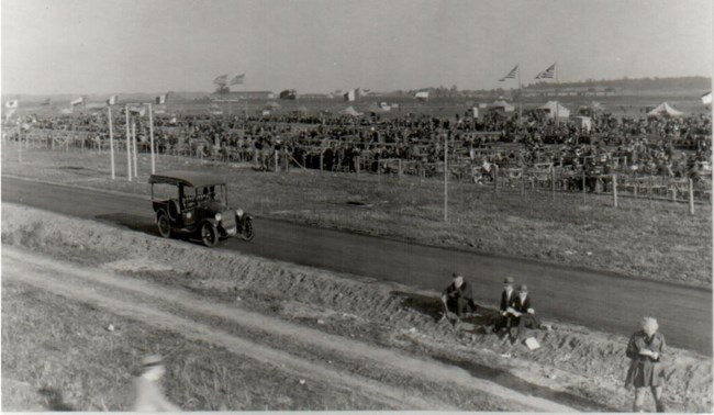 Spectator Stands Black and white image of a grassy airfield with stands set up. A large amount of individuals are seated in the spectator stands. An old car sits in the left side of the image on a paved road. A few people appear in the lower right of the image.