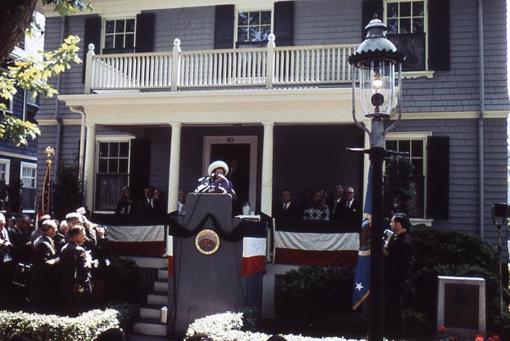 Mrs. Rose Kennedy speaks at a podium in front of a grey 2 story house.