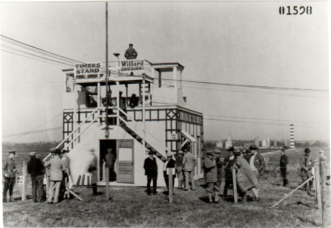 Timers Stand for Air Races Black and white image of a timers stand set up on a grassy field. There are many individuals standing in the foreground of the image in front of the stand. There are wires and a pole near the stand.