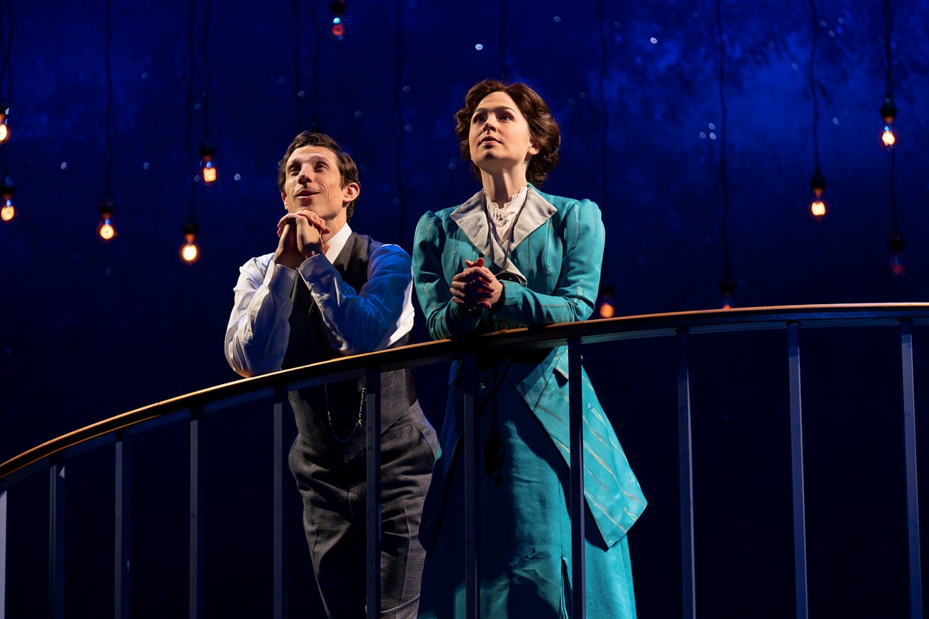 A man and a woman stand leaning on a railing in front of a starry night background with hanging lights.