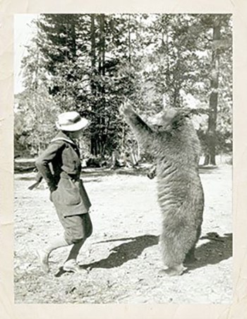 A woman wearing breeches with her hands clasped behind her back dances with a bear.