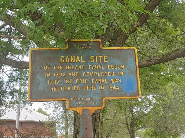 A rusted NYS historic marker. "CANAL SITE of the Inland Canal begun in 1792 and completed in 1797. The Erie Canal was relocated here in 1844."