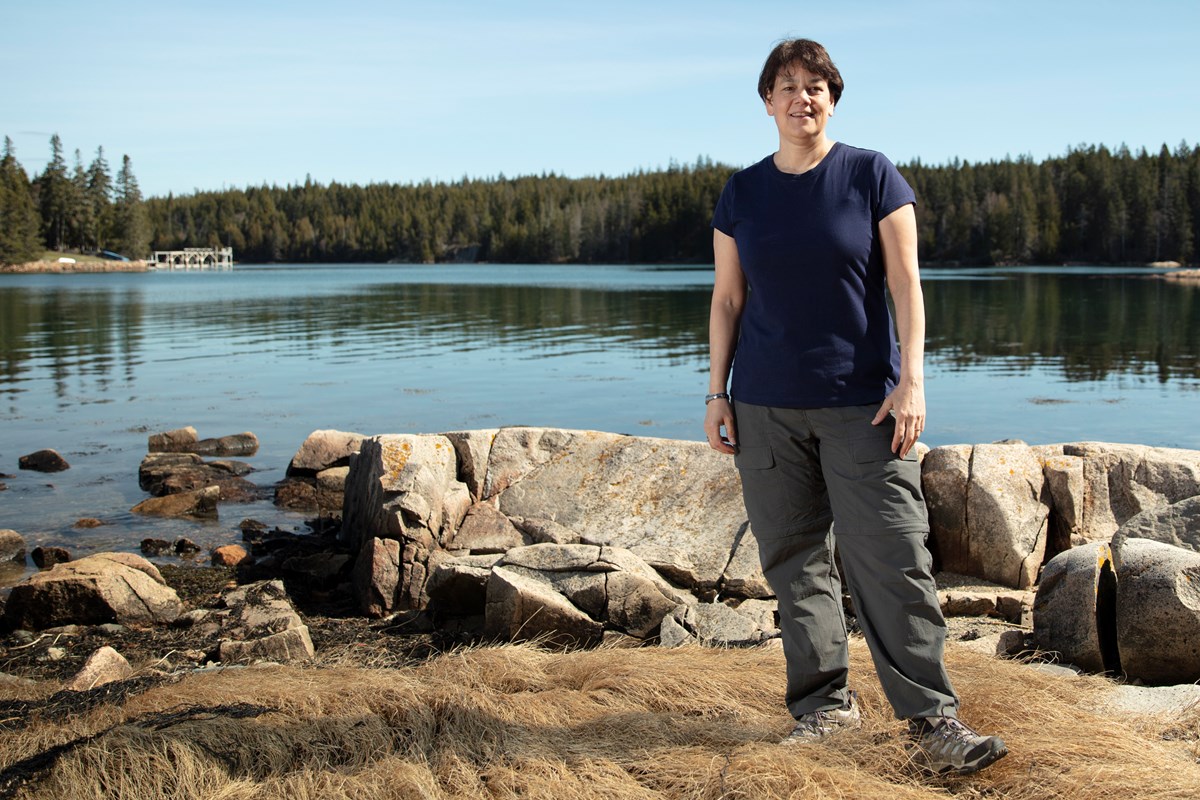 Woman with short, dark brown hair, wearing a dark blue shirt, stands in front of large rocks on a lake shore. Evergreen trees are in the distance.