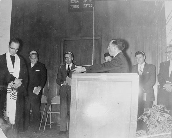 Rabbi Sanders Tofield Rabbi Sanders Tofield speaking to six men surrounding him and listening.