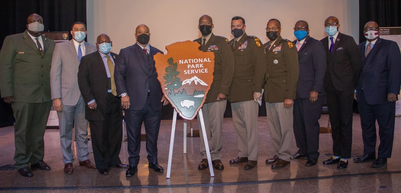 Nine men stand shoulder to shoulder looking at the camera. Three of them are wearing military uniforms and the other six are wearing suits.