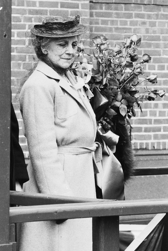 Woman in a dress suit and hat standing next to the bow of a ship.