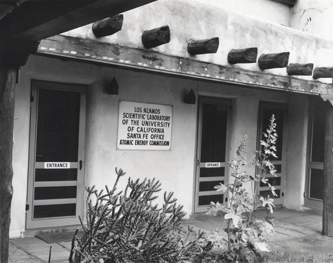 The entrance of a flat-roofed adobe building with 2 screen doors and a sign that reads Los Alamos Scientific Laboratory of the University of California Office.