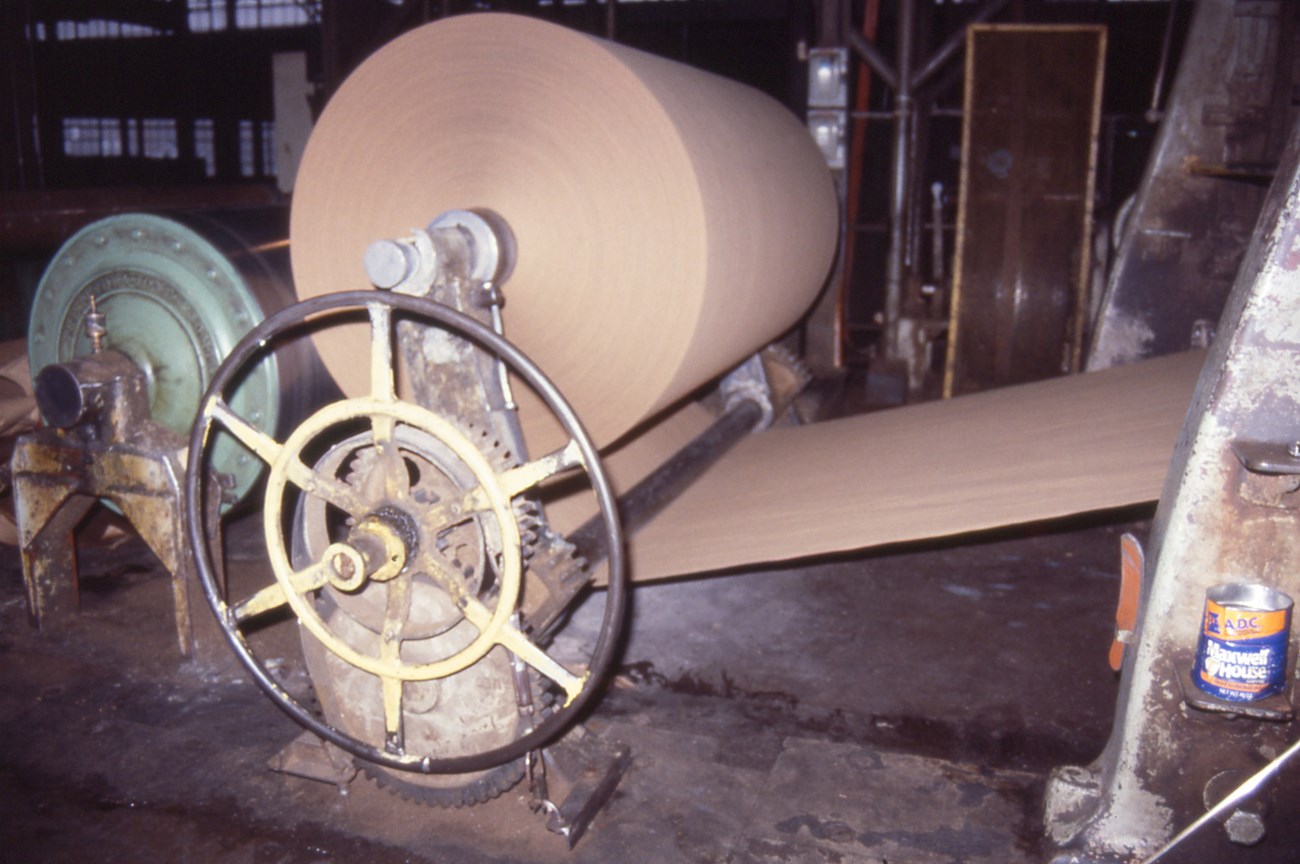 A large roll of beige paper spins on metal machinery inside a factory.