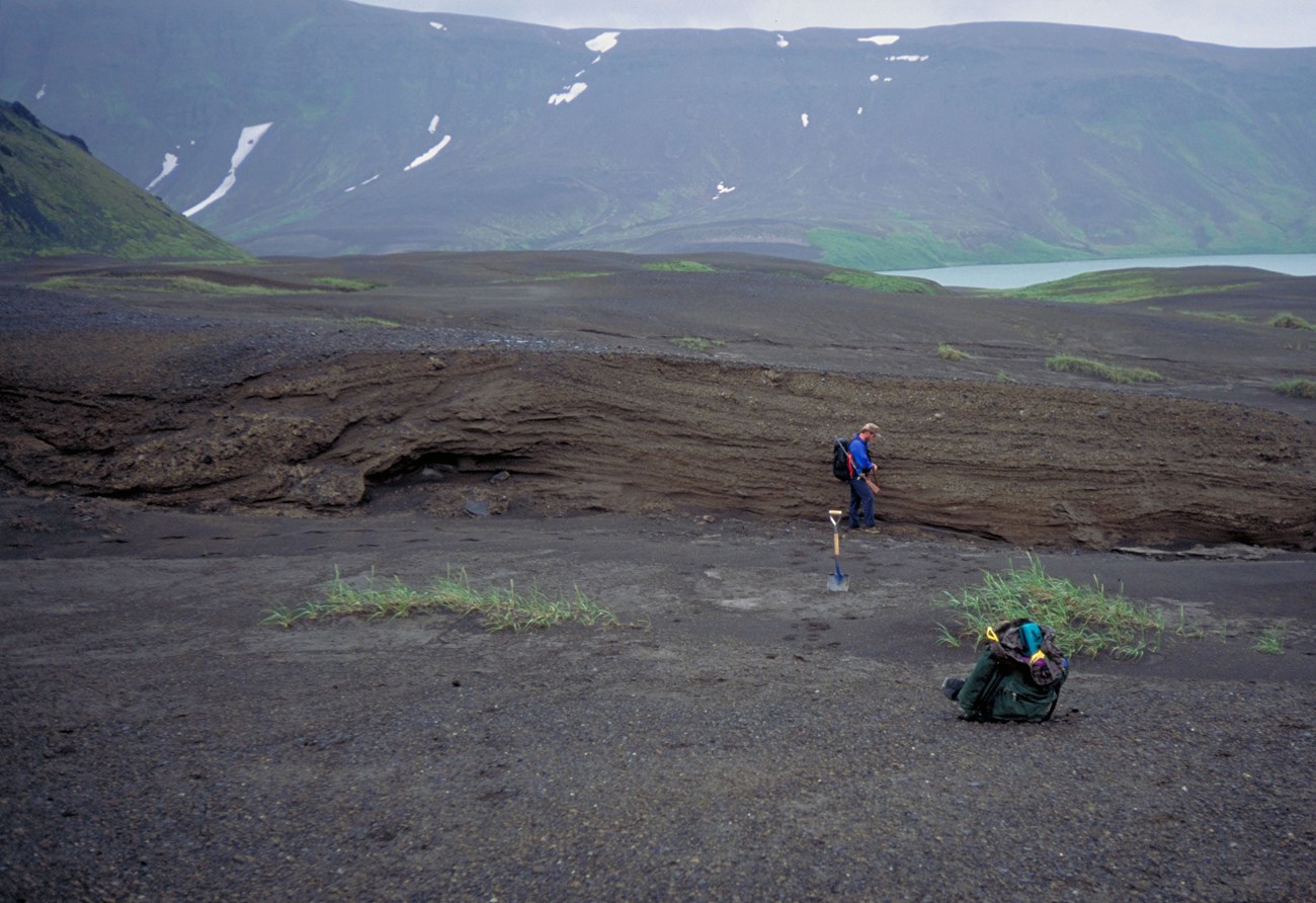 Photo of a volcanic landscape with a person standing in the middle and a lake and mountains in the distance.