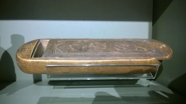 1192 A small wooden rectangular box, with rounded ends and a simple carved ornamental design, sits behind glass in a museum display. The sliding cover is partially open, showing the inside of the case, which is large enough to hold a shaving razor.