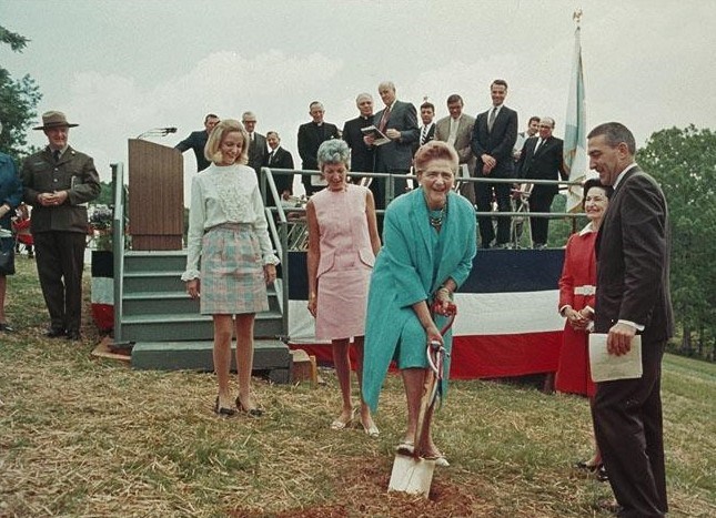 Color photo of a woman in a bright dress suit pushing a golden shovel into the ground while a nearby crowd of mostly men in suits looks on.