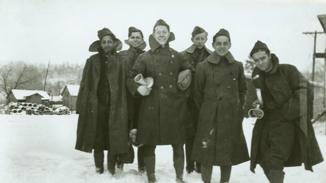 Six young men in winter dress coats and military style hats smile for the camera and hold mess kits; snow covers the ground and a building behind them.