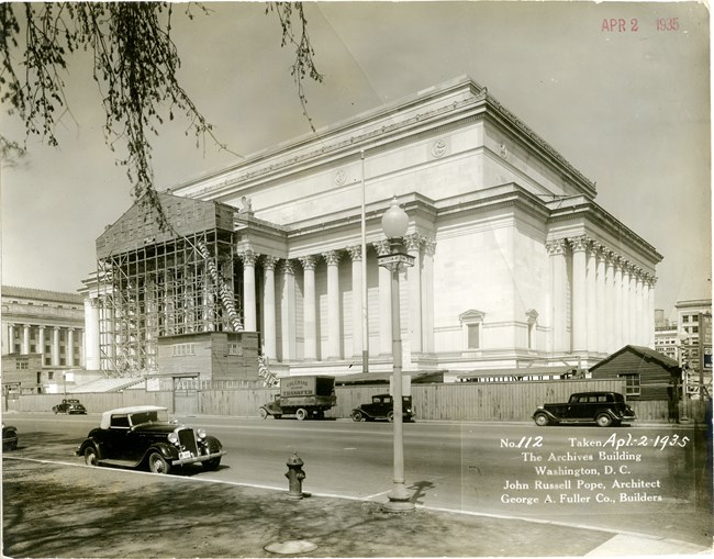 Black and white image showing a large classical building styled after Roman architecture. The building is under construction. The left side has scaffolding covering the pediment and supporting columns. 1930s cars are parked streetside in the foreground.