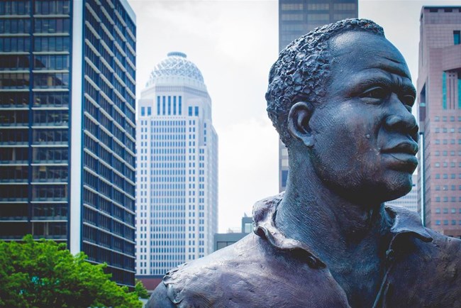 A close-up of the York statue.  Seen is York’s head looking to the right. In the background is the skyline of downtown Louisville, Kentucky.