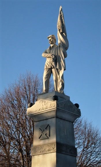 Tall stone monument, with soldier standing atop pedestal, holding a flag, tree, no leaves, in background
