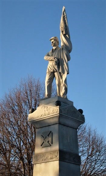 128thmonumentham Tall stone monument, with soldier standing atop pedestal, holding a flag, tree, no leaves, in background