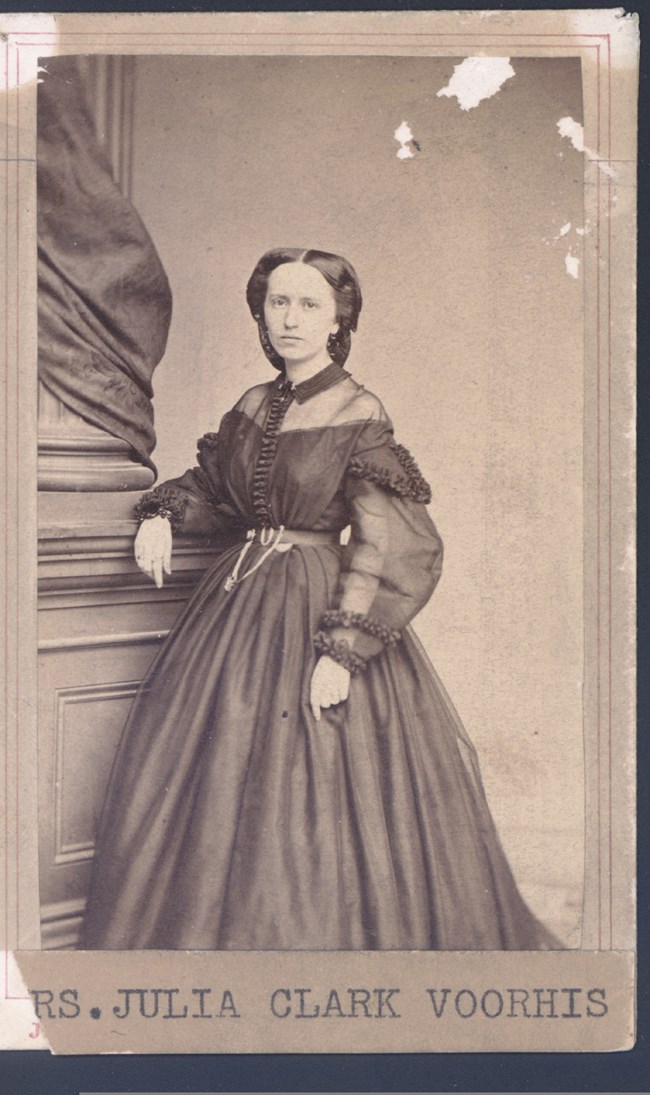 1298 An old black and white photo shows Julia Clark Voorhis standing, leaning against a large column. She is wearing a dark-colored dress of the period.