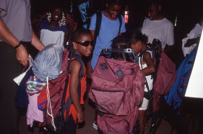 A group of Black boys and girls try on backpacks full of camping gear.