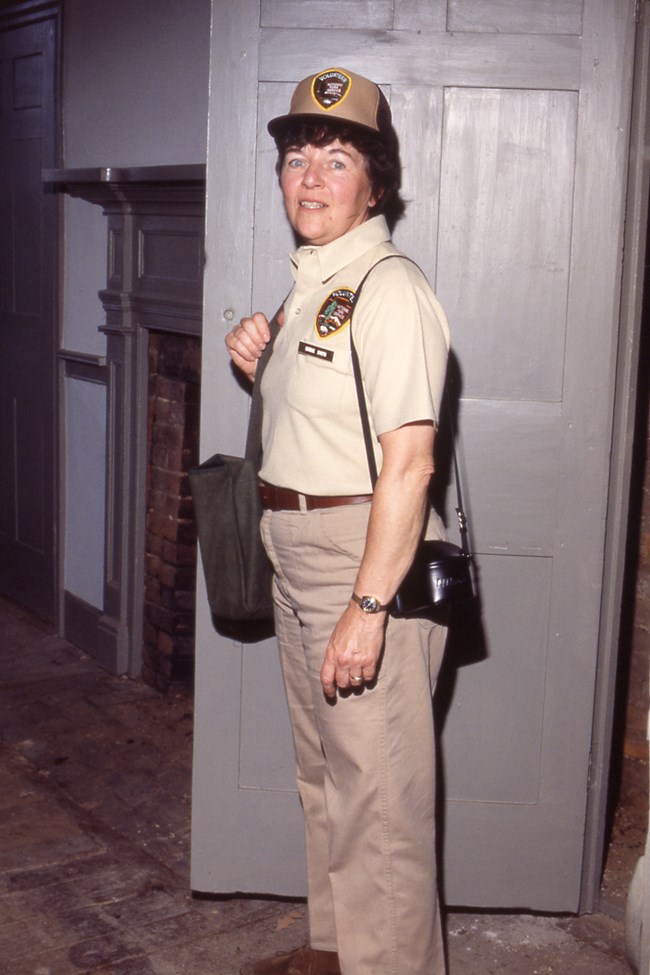 A woman in a beige uniform with volunteer patches stands inside a historic building with a satchel and a camera hanging from her shoulders.