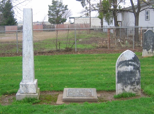 A tall, thin, white column grave stone of Alexander Hamilton Willard is on the left, a flat bronze marker is in the middle, and the marker for Willard’s wife is on the right.