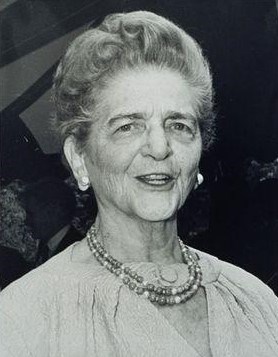 Black and white head and shoulders photograph of an older white woman. She is smiling, has short light-colored hair, and sports round earrings and a short double-stranded necklace