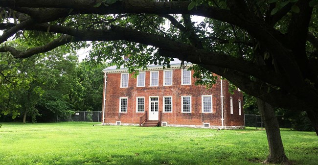 1360 The Jarrot Mansion, a two-story red brick home. On the first floor, the entry door is in the center, with two large windows on the left of the door and three windows on the right. Six large windows span the entire second floor. A large tree frames the