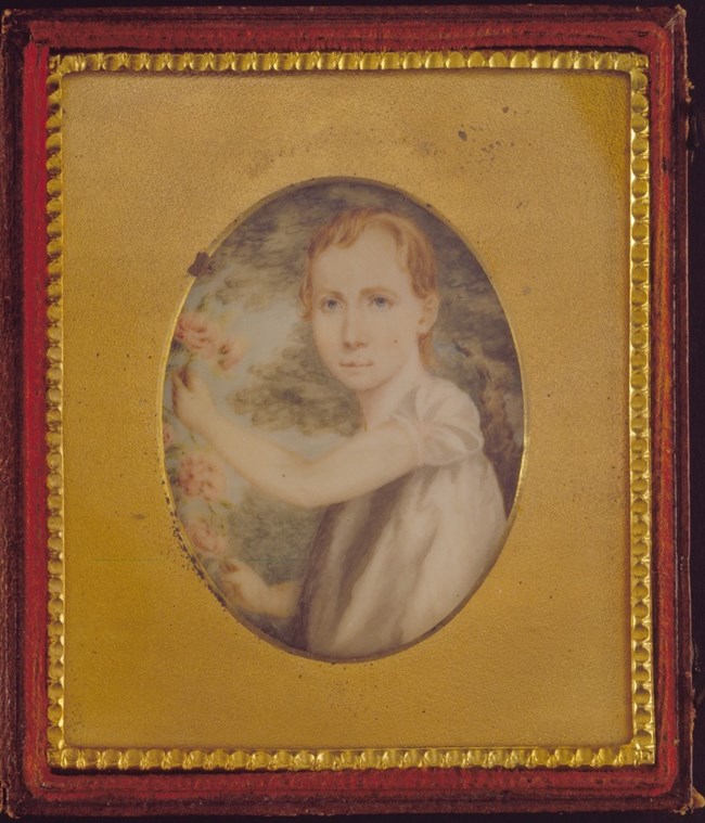 An oval portrait of Mary Margaret Clark.  She is looking at the viewer, with blue eyes and short auburn hair.  She is wearing a short-sleeved blouse, holding a pink rose in her left hand.  The portrait is within a gold-colored frame surrounded by a burgun