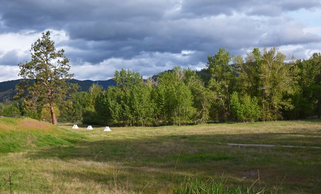 1380 A grassy area at Travelers’ Rest State Park, surrounded by cottonwood trees. Three small white tents can be seen near a large pine tree. Mountains and heavy clouds are in the background.