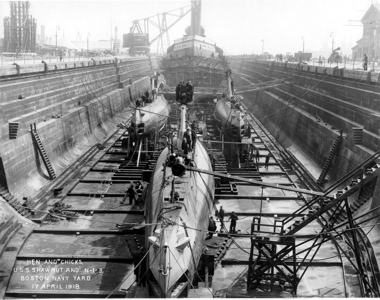 A dry dock in a shipyard with submarines in the foreground and a minelayer ship in the background.