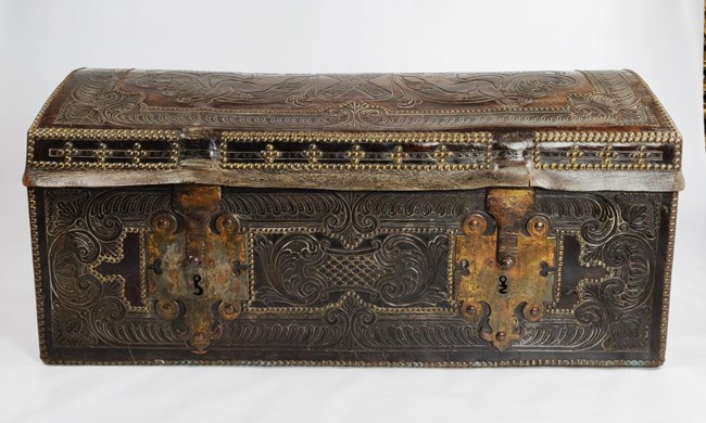 1425 A dark rectangular trunk, with ornate designs in leather. Two large metal latches are on the front.