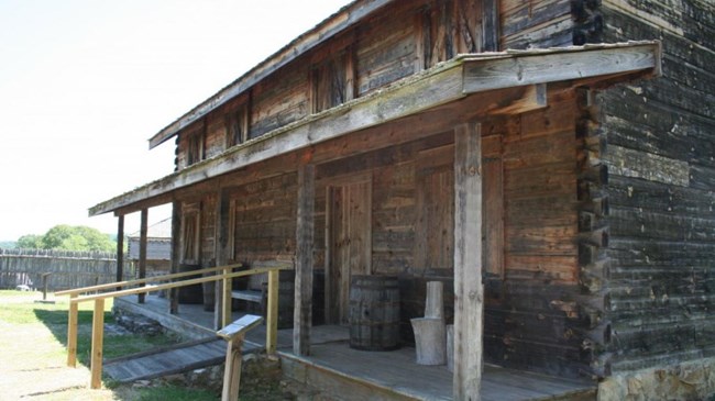 1434.0 A rough-hewn cabin with a covered porch is in the foreground. Behind it is the fort’s wooden palisade with a corner guard house.