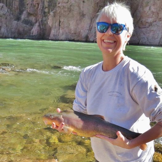 a woman with blond hair and sunglasses is smiling while holding a large fish. A flowing river is in the background.