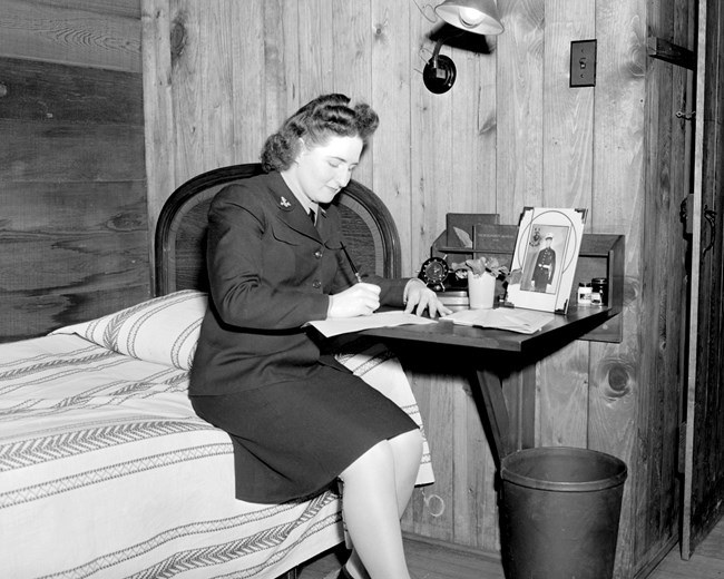 WAVE in Sugar Camp cabin - May 19, 1943 Woman wearing dark, uniform dress writes a letter on her bedside table while sitting on the edge of her bed.