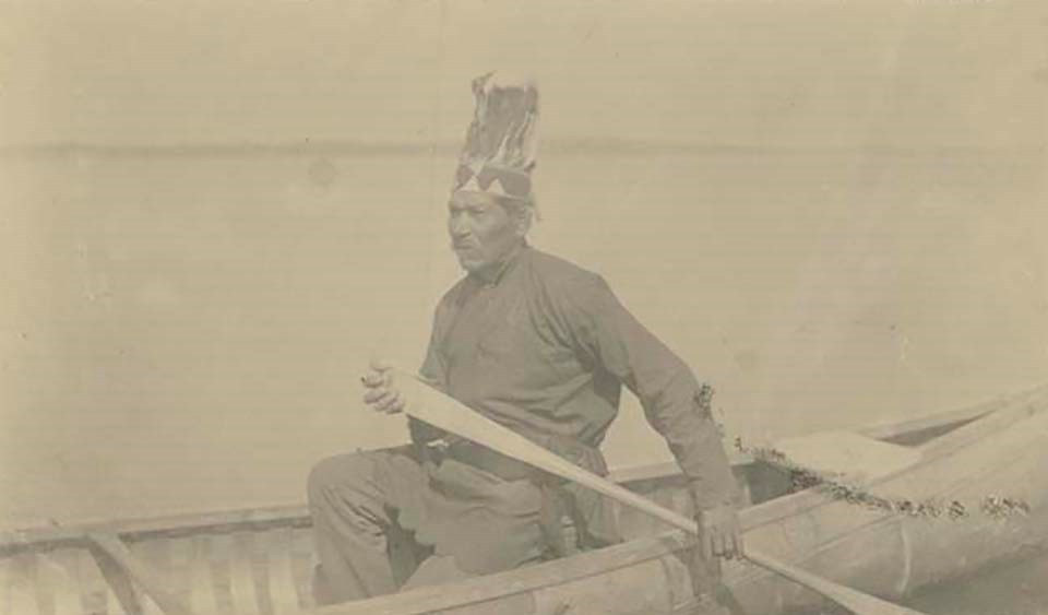 An elderly man with feathered head dress paddles a canoe in open water.