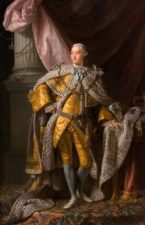 King George III an adult white man with white powdered hair. He is wearing a gold outfit, resplendent with white royal robes. He is adorned with light blue bows and jewels. The background is lavish, with a pillar, curtain, and patterned rug. The royal crown sits to the l