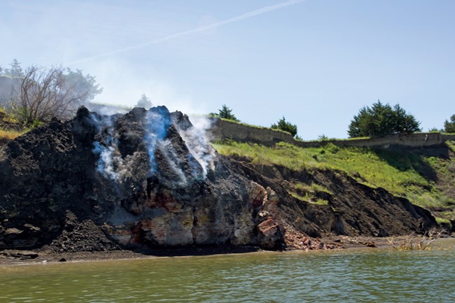 A photograph shows a small, dark rock face along the Missouri River which has smoke rising from it.  Around the rock are green grass embankments with a few scattered trees.  The river is in the immediate foreground.  