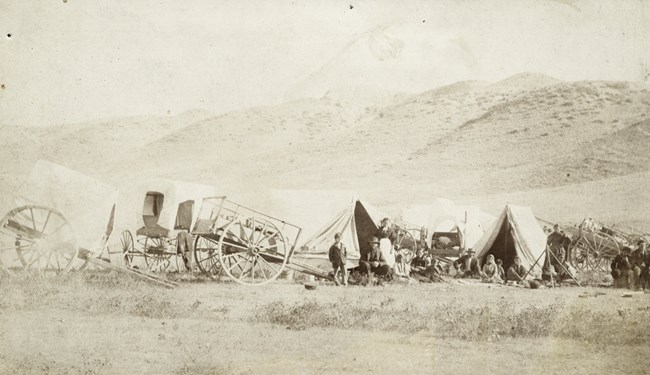 Black and white photograph of several tents and carts. A group of people stand in front of the small camp and look towards the camera; behind them and surrounding them are tall grasses.