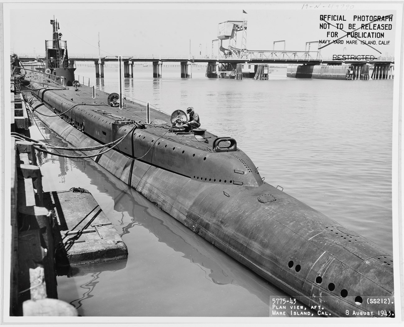 Long, narrow submarine partially above water, at a dock, with a person sitting by a top hatch.