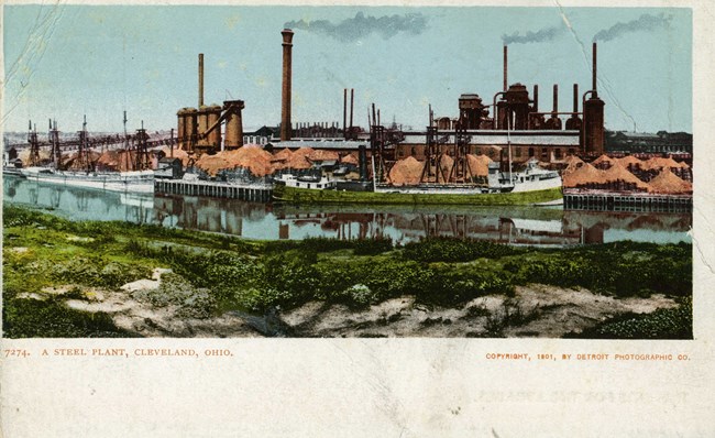 A colorized black-and-white photo of a steel plant's smoking towers with the river in the foreground.