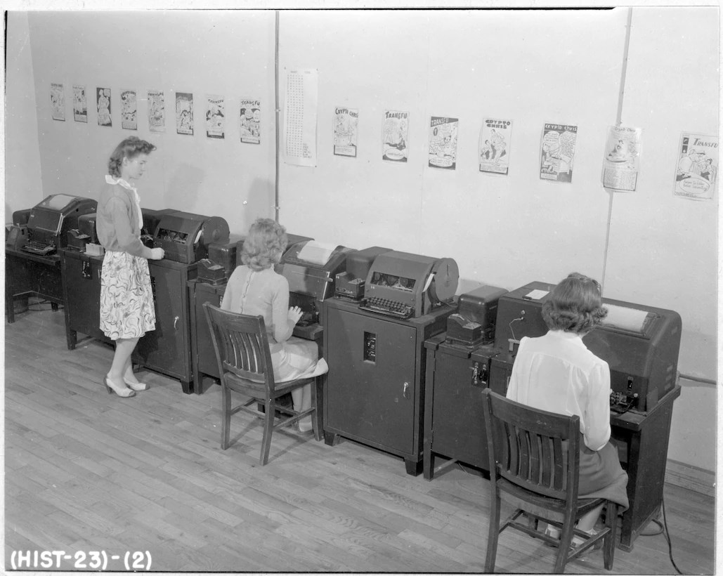 Black and white photo of two women sitting and one standing at desks facing the wall. There are posters hung along the wall they are facing.