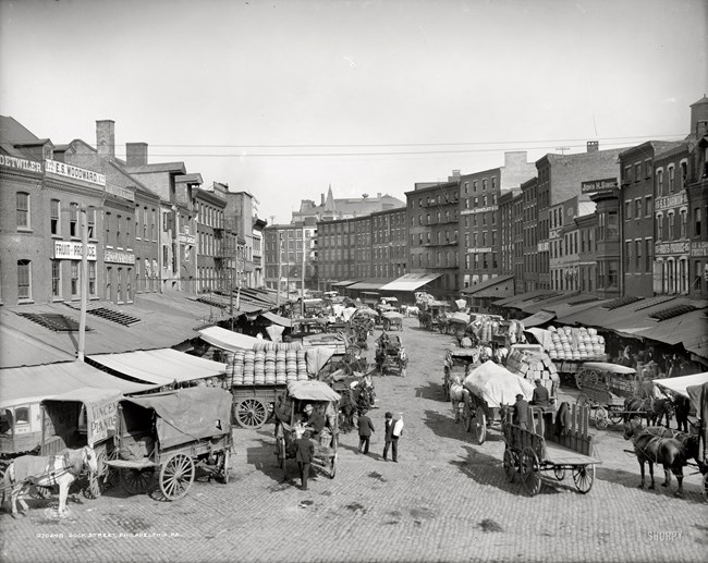 Historic urban market, busy with vendors and their horses and wagons filled with goods.