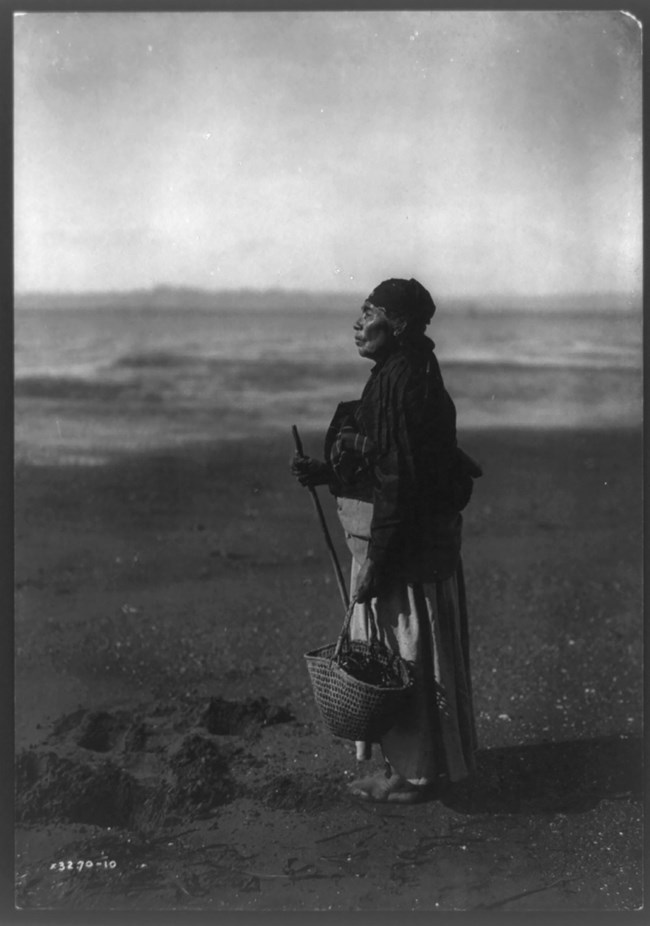 Black and white photo of a Chinook woman on a beach. She is standing facing the water in a long dress and head covering, holding a basket.