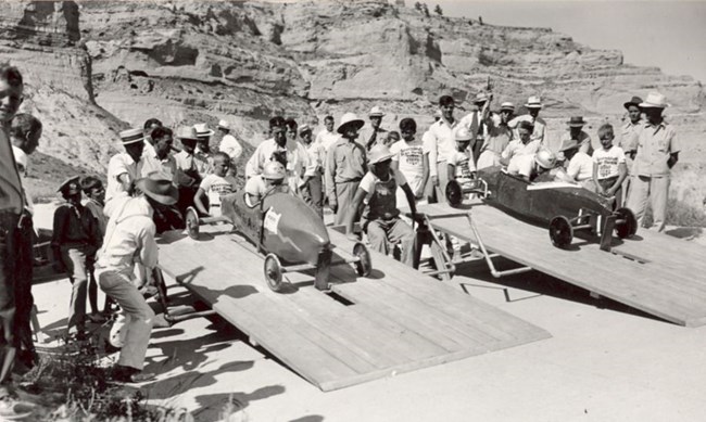 A black and white photo of two soapbox cars on ramps with people gathered around.