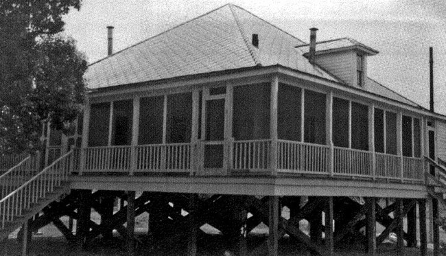 A 20th century cottage on stilts with a wraparound porch is pictured in black and white with foliage in the left side background