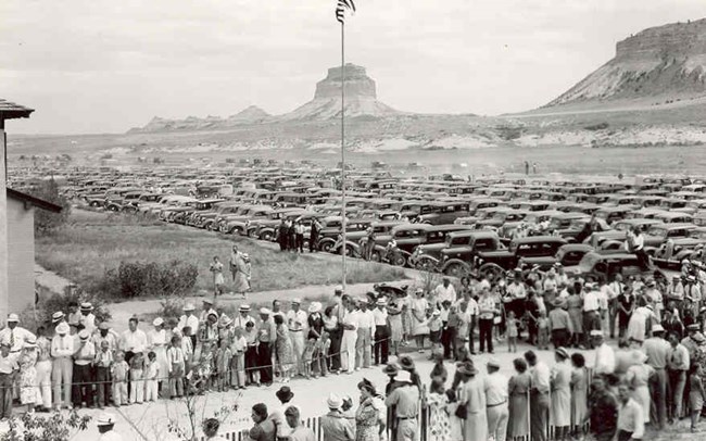 Crowds of people line a road with rows of automobiles in the background.