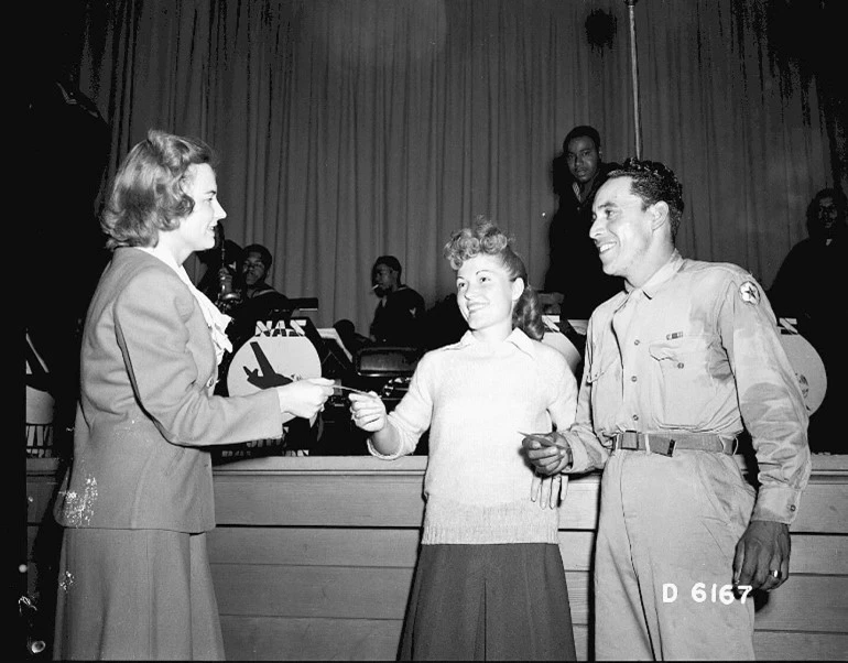 A man and woman stand side by side facing a woman who is handing them an award. A 5-person band is in the background.
