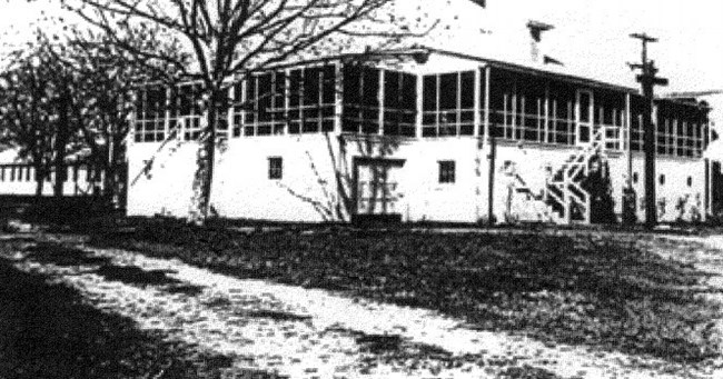 A white 20th century cottage house with a wraparound porch is pictured in black and white. A path and a few leafless trees can be seen on the left side.